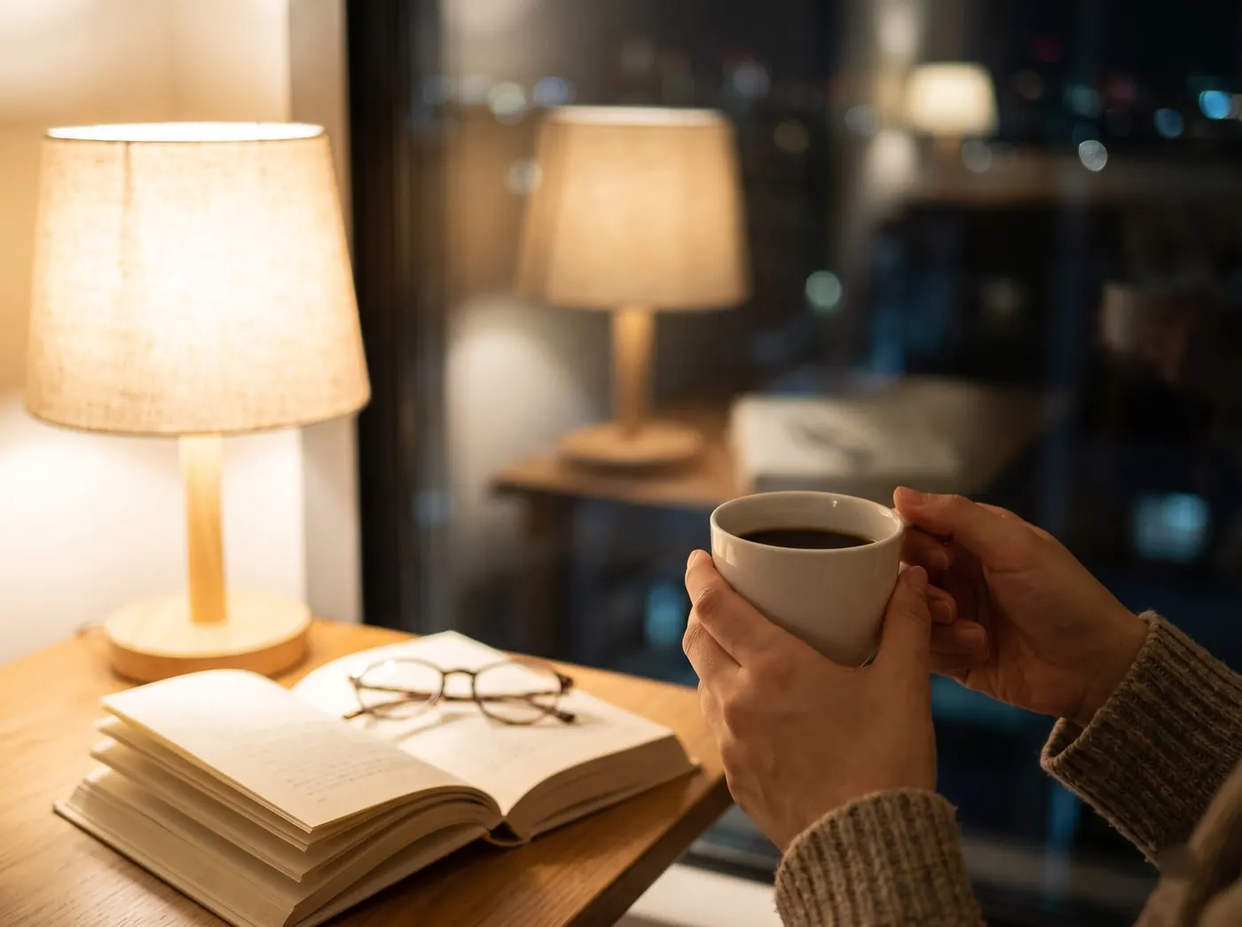 Hands holding a cup of coffee beside a lamp and an open book