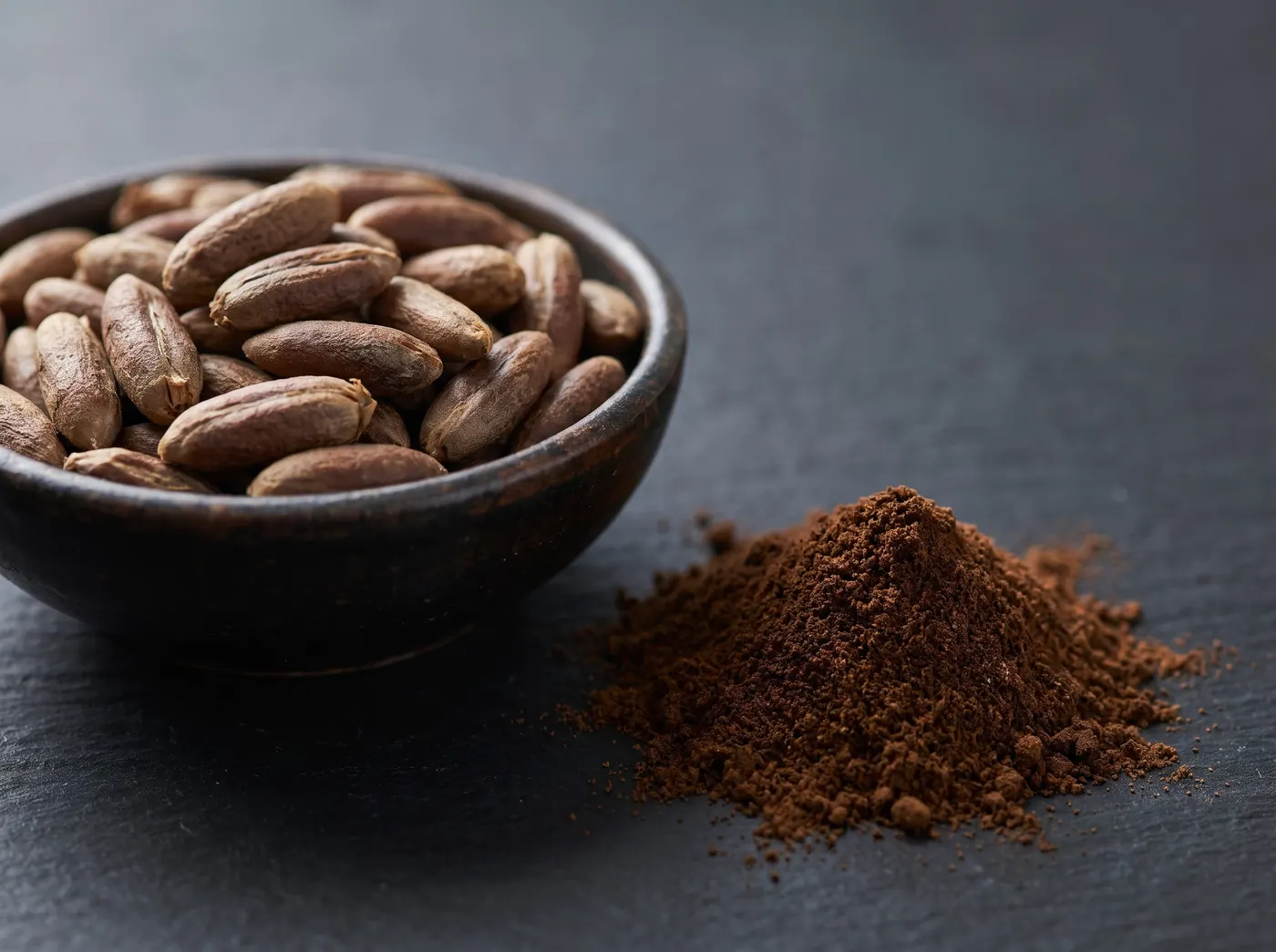Roasted date seeds in a dark bowl beside a pile of finely ground date seed powder